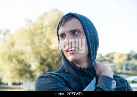 Junge attraktive blauäugige Mann lächelt im Freien, trägt Hoodie Blick zur Seite an einem sonnigen Herbst oder Frühlingstag auf dem Hintergrund der Natur Stockfoto