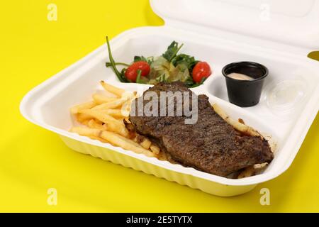 Gegrilltes Steak mit pommes Frites, Gemüse und Sauce in einem Kunststoffbehälter isoliert auf einem gelben Hintergrund. Lieferung von Lebensmitteln. Stockfoto