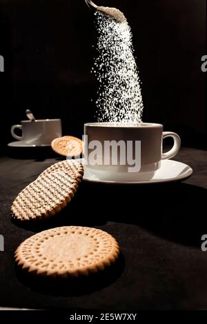 Anordnung von weißen Tassen, mehreren Keksen und schwebendem Zuckerwürfel. Stockfoto