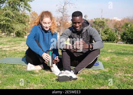 Afrikanischer schwarzer Mann und Rotschopf kaukasische Frau überprüfen das Smartphone nach dem Training in einem Park mit einer Matte. Fitness und Sport im Freien. Stockfoto