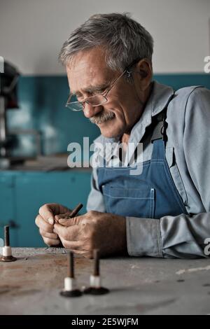 Ein älterer Meister mit Brille bei der Arbeit. Ein weißer alter Mann mit kaukasischem Aussehen sitzt an einem Tisch und arbeitet mit seinen Händen Stockfoto