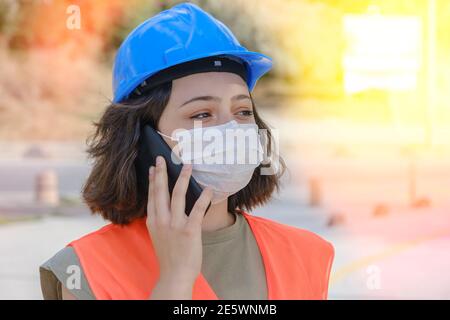 Junge Frau mit blauem Helm und Gesichtsmaske im Gespräch auf dem Handy auf der Baustelle. Neue Normalität während der Coronavirus-Pandemie. Stockfoto