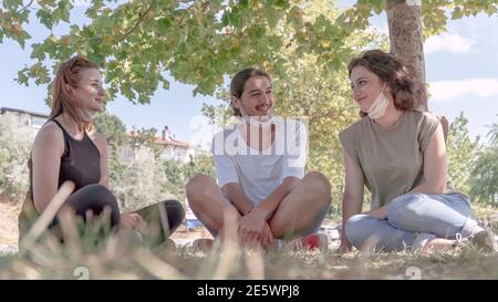 Junge Freunde reden und lachen, während sie auf Gras sitzen. Treffen mit Freunden im Park während der Coronavirus-Epidemie. Stockfoto