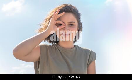 Charmante junge Frau tut ok Geste mit ihrer Hand und Blick durch OK Zeichen lächelnd mit glücklichen Gesicht vor Himmel Hintergrund. Gesichtsausdruck. Stockfoto