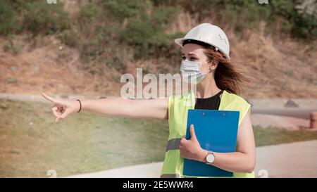 Junge Bauarbeiterin mit weißem Helm und Gesichtsmaske, die mit der Hand auf die Baustelle zeigt. Stockfoto