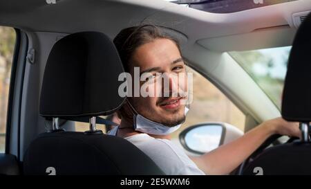 Junger, hübscher Mann in Maske sitzt auf dem Beifahrersitz in einem Auto lächelt und schaut auf die Kamera. Stockfoto