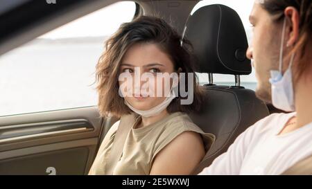 Glückliches junges Paar, Freunde mit Gesichtsmasken sitzen im Auto und reden sich gegenseitig. Roadtrip in neuer Normalität. Corona-Virus-Pandemiekonzept. Stockfoto