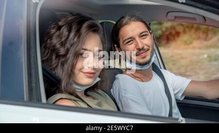 Glückliches junges Paar, Freunde mit Gesichtsmasken sitzen im Auto und Blick auf die Kamera. Roadtrip in neuer Normalität. Corona-Virus-Pandemiekonzept. Stockfoto