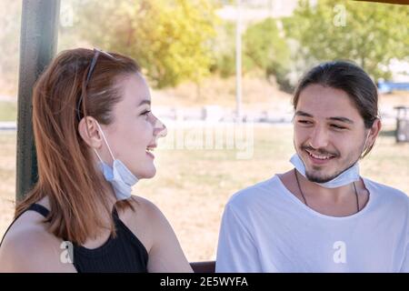 Glückliche junge Freunde tragen Gesichtsmasken bei einem Gespräch im Park während der Coronavirus Ausbruch. Stockfoto