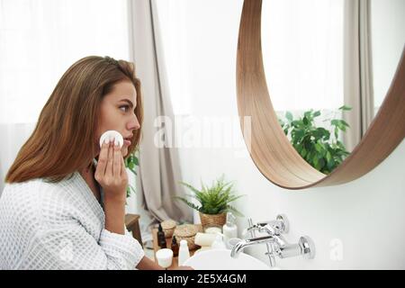 Frau mit einem Schwamm Wattepad Problem Haut. Ein Bild von einem glücklichen Mädchen, das ihr Gesicht mit Wattebäcken über dem Badezimmer putzt. Schönes Gesicht der jungen Frau Stockfoto