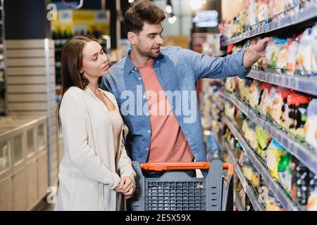 Junges Paar, das in der Nähe von Einkaufswagen und Regalen mit Lebensmitteln steht Auf verschwommenem Vordergrund im Supermarkt Stockfoto