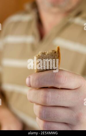 Ein Mann in einem gestreiften Hemd reicht der Kamera einen Laib Brot. Ein kleines Stück Roggenbrot mit gezackten Rändern. Barmherzigkeit, Spende, Großzügigkeit. Frontpartie Stockfoto