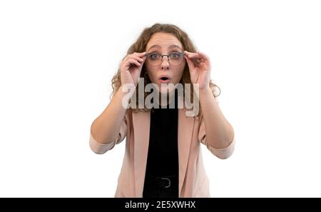 Junge überraschte Frau hält ihre Brille mit den Händen. Sie trägt einen korallenfarbenen Anzug, isoliert auf weißem Hintergrund. Stockfoto