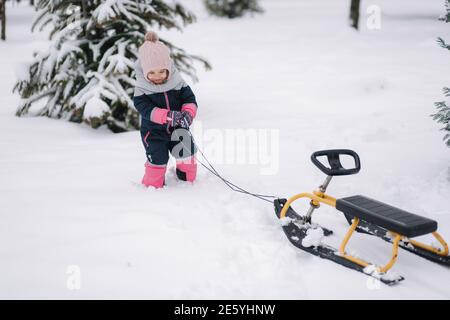 Kleines Mädchen, das auf Schnee läuft und ihre Schlitten hochzieht. Aktive Freizeit im Freien im Winter. Nettes kleines Mädchen Stockfoto