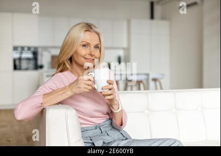 Bezaubernde Frau mittleren Alters sitzt auf der Couch und entspannt mit einer Tasse Kaffee. Ein Porträt einer charmanten blonden Frau, die zu Hause träumt Stockfoto