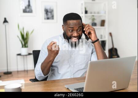 Erfolgreicher afroamerikanischer Freiberufler im Telefongespräch, glücklich, das Ziel zu erreichen. Junger Geschäftsmann, der auf den Bildschirm des Laptops schaut, Gewinner Stockfoto