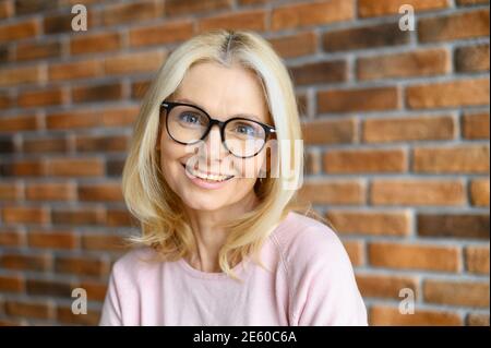 Elegante Geschäftsfrau mittleren Alters trägt stilvolle Brille schaut auf Kamera und lächelt. Nahaufnahme Porträt der charmanten blonden Frau mit einer Backsteinmauer an Stockfoto