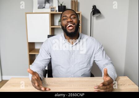 Aufgeregt junger afroamerikanischer Arbeiter, der mit weit geöffneten Händen am Schreibtisch sitzt und die Kamera anschaut, legte die Hände auf einen Tisch, erfolgreich Stockfoto
