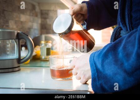 Nahaufnahme Mann im Hausbademantel Gießen heißen Tee aus Glas Teekanne in Tasse auf dem Küchentisch. Bereiten Sie morgens Tee zu. Eine Tasse frisch Stockfoto