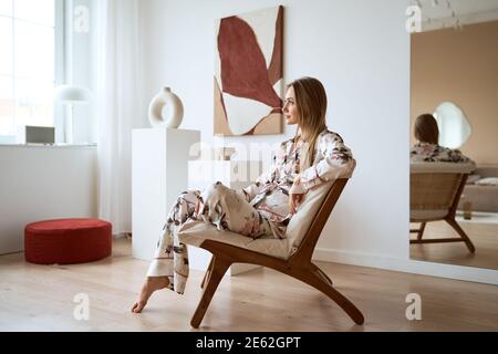 Junge Frau in Seide Pyjamas ruhen und sitzen auf bequem Stuhl zu Hause Stockfoto