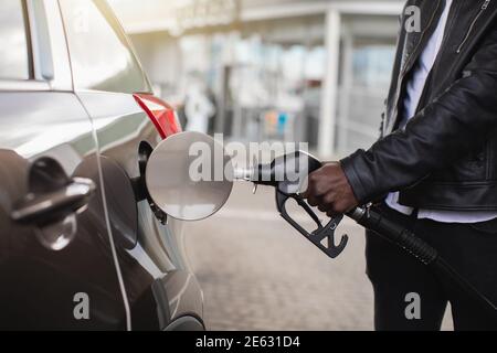 Beschnittenes Nahaufnahme Bild von unkenntlichen afrikanischen Geschäftsmann in Casual Wear, halten in der Hand Füllen Pistole und tanken sein Luxus-Auto mit Benzin Stockfoto