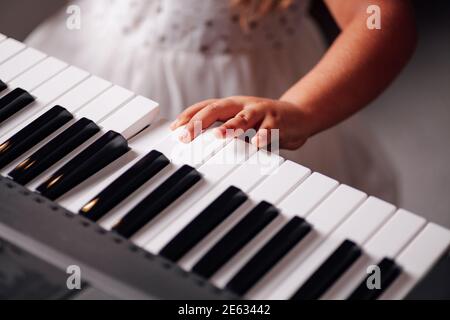 Nahaufnahme, drückt die Hand eines fünfjährigen Mädchens in einem weißen Kleid ihre Finger auf die Tasten eines elektronischen Synthesizers im Wohnzimmer Stockfoto