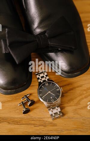Schwarze Fliege an Herrenschuhen mit Armbanduhr und Manschettenknöpfen auf Holzstruktur. Stockfoto