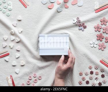 Hand halten Leuchtkasten mit Copy-Space. Flaches Lay mit Leuchtkasten auf Elfenbein, aus weißem Textil. Herzen, Süßigkeiten, Knöpfe, verschiedene Dekorationen auf angeordnet Stockfoto