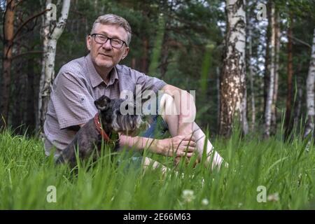 Ein Mann mittleren Alters in Brille sitzt mit seinem Hund auf dem Gras im Park vor dem Hintergrund von Bäumen. Ruhen Sie sich nach einem langen und angenehmen Spaziergang aus. Stockfoto