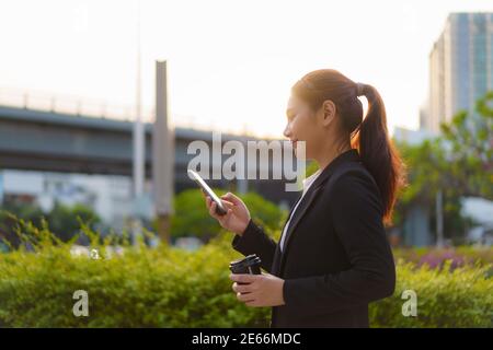 Asian Executive working woman Holding Coffee Cup und mit einem Handy in der Straße mit Bürogebäuden im Hintergrund in Bangkok, Thailand. Stockfoto