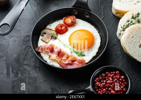 Spiegeleier mit Speck und Gemüse in gusseiserner Bratpfanne, auf schwarzem Hintergrund Stockfoto