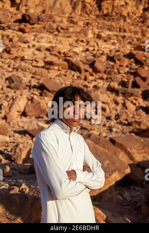 Wadi Rum, Jordanien 03.31.2010: Ein junger beduinenmensch in einem langen weißen jubba-Kleid, der in der Wadi Rum Desert mit seinen Armen von einem Felsen gekreuzt steht. Stockfoto