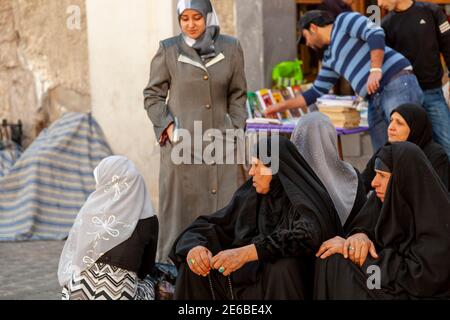 Damaskus, Syrien 03-28-2010: Eine Gruppe älterer muslimischer Frauen mit schwarzen Burkas sitzt auf dem Bürgersteig beim Basar. Andere Frauen mit bunten Stockfoto