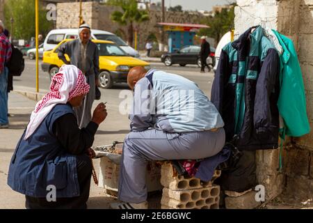 Hama, Syrien 04-02-2010: Zwei syrische Männer sprechen auf der Straße. Einer sitzt auf einem Ziegel und versucht, Perlen und kleine Gegenstände auf seinem kleinen Display zu verkaufen. Stockfoto