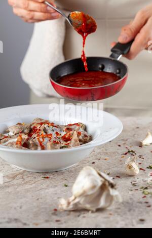 Eine Küchenchefin trägt Schürze fügt frische Tomatensauce zu hausgemachten türkischen Knödel in Joghurt. Dieses traditionelle Gericht ist als Manti bekannt und wird serviert Stockfoto