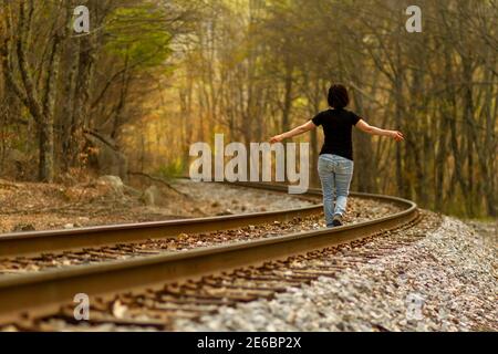 Eine junge kaukasische Frau mit Jeans und T-Shirt geht mit weit geöffneten Armen auf den Bahngleisen an einem ländlichen Ort. Ein stimmungsvolle Konzept für f Stockfoto