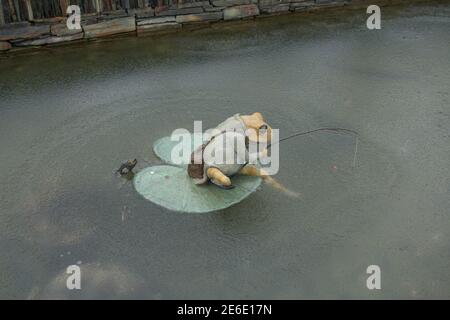 Keramik Frosch sitzt auf einem Lily Pad mit einer Angelrute in einem gefrorenen Teich im Rosemoor Garten in Rural Devon, England, Großbritannien Stockfoto