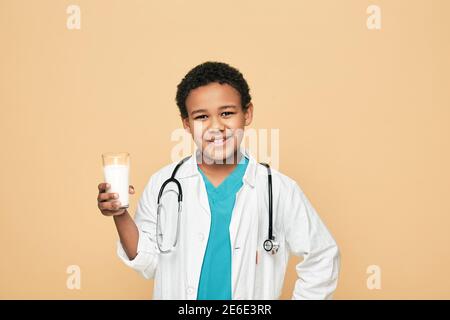 Afroamerikanischer Junge, gekleidet wie ein Arzt, hält ein Glas Milch auf beigem Hintergrund. Vorteile von Milch für Kinder Stockfoto