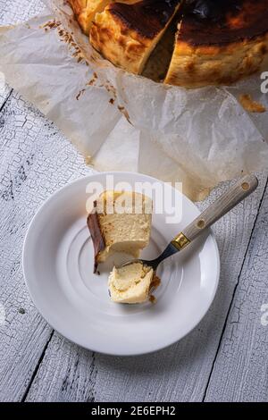 Bissen Sie einen Teil des baskischen verbrannten Käsekuchen auf einem weißen Teller mit Vintage-Löffel an der Seite und teilweise sichtbar geschnittenem Käsekuchen auf einem weißen Holztisch. Hausgemacht Stockfoto