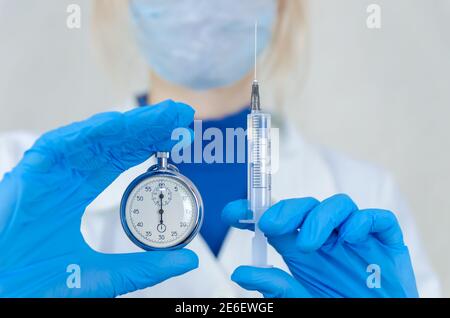 Maskierte Ärztin hält eine Spritze mit einer Nadel und einer Stoppuhr vor sich. Konzept der Zeit in der Medizin, Impfung Stockfoto