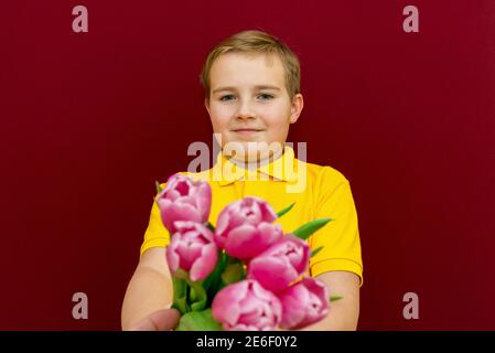 Junge geben Blumen Bouquet, Kind hält lila Tulpen Bündel, Blick auf Camera.Studio rot oder burgunderrot Hintergrund. Stockfoto