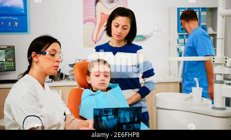 Pädiatrischer Zahnarzt zeigt zahnärztliche Probleme halten Radiographie zeigt auf betroffene Zähne, während Mann Assistent Vorbereitung sterilisierte Werkzeuge für die Operation. Arzt und Krankenschwester arbeiten in stomatologie Einheit Stockfoto