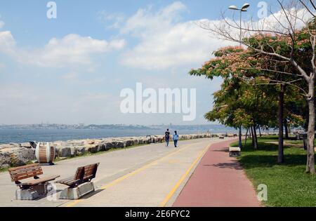 Ein paar Leute, die durch einen Park am Wasser in Moda, Istanbul mit der alten Skyline in der Ferne laufen. Stockfoto