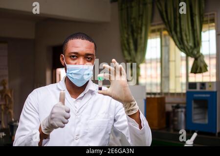 Junger schwarzer Laborwissenschaftler, der eine Nasenmaske trägt und eine Blutprobe in der Hand beobachtet Stockfoto