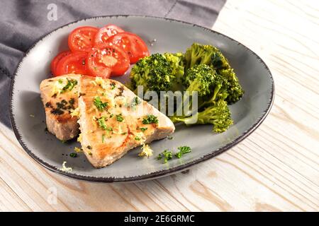 Gebratener Thunfisch Scheiben mit Brokkoli und Tomaten auf einem asymmetrischen grauen Teller und einem leichten Holztisch, gesunde Mahlzeit zur Gewichtsreduktion mit ketogenen oder l Stockfoto