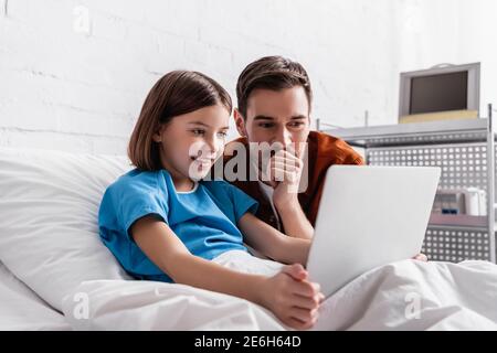 Fröhliches Mädchen mit Laptop im Krankenhausbett in der Nähe glücklicher Vater Stockfoto