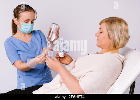 Ein älterer Patient liegt auf einem Stuhl und ein junger Arzt bereitet Vernebler für die Behandlung von Erkältungen und Viren Atemwegserkrankungen auf weißem Hintergrund Stockfoto