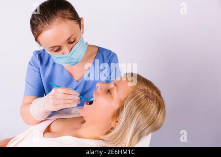 Der junge Zahnarzt in einer medizinischen Maske überprüft den Zustand der Zähne einer älteren Frau, die auf einem Stuhl auf einem grauen Hintergrund mit Seitenraum sitzt. Stockfoto