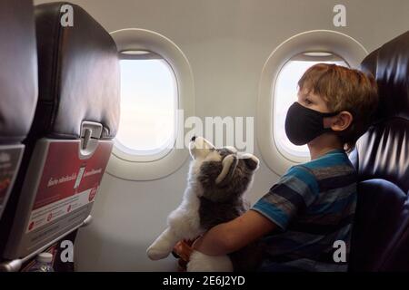 Ein Kind in einem Flugzeug, das ein weiches Spielzeug hält und eine Gesichtsmaske trägt. Stockfoto