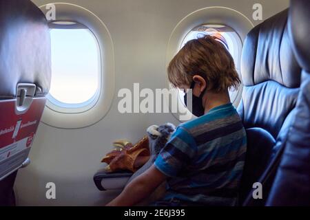 Ein Kind in einem Flugzeug, das ein weiches Spielzeug hält und eine Gesichtsmaske trägt. Stockfoto